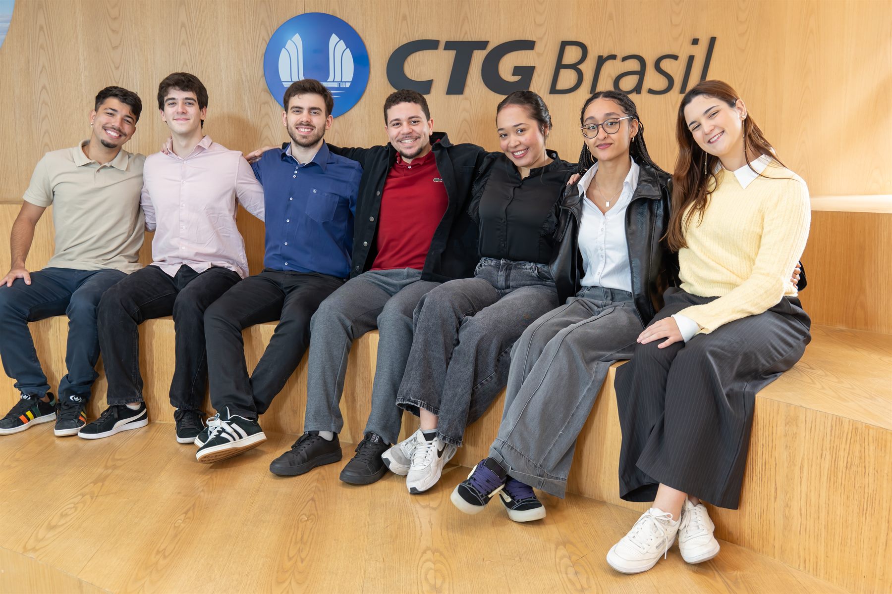 Turma de estagiários da CTG Brasil sentada na arquibancada de madeira, em frente ao logo da empresa